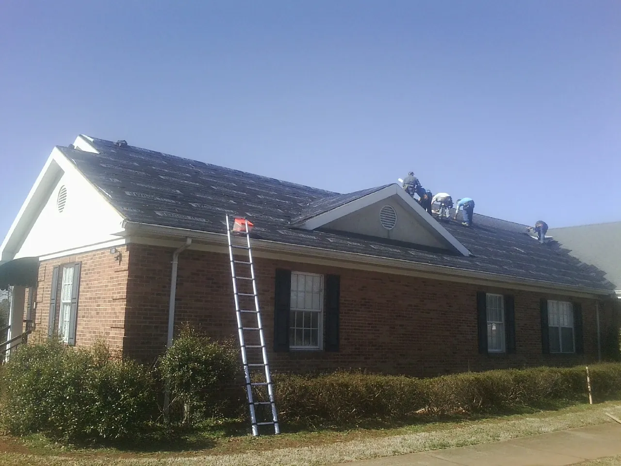 Crew tearing shingles off a brick ranch roof with a ladder set — Metal Roof Repair work by our crew in Elberon
