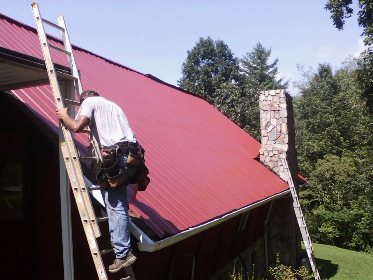 Roofer on a ladder beside a red metal roof and stone chimney — Soffit & Fascia Repair by Roofing Brothers in Elberon